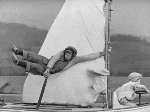 A monkey hangs on to a mast while a baby steers a sailboat in a black and white film