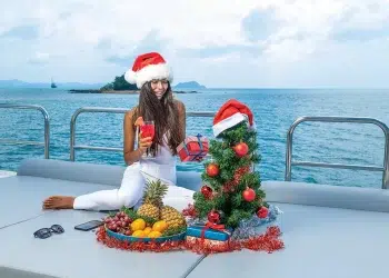 A woman with a Santa hat opens presents in front of a small Christmas tree on a boat
