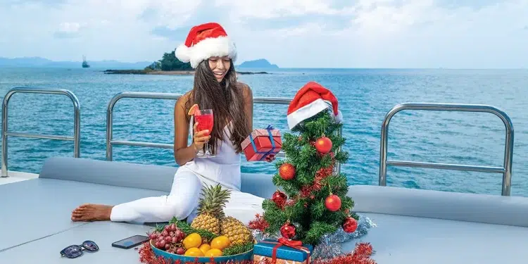 A woman with a Santa hat opens presents in front of a small Christmas tree on a boat
