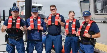 5 US Coast Guard members stand infront of a boat wearing their life jackets (Personal Floatation Devices)