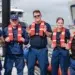 5 US Coast Guard members stand infront of a boat wearing their life jackets (Personal Floatation Devices)